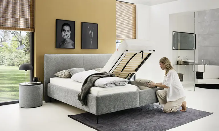 Graues Bett mit angehobenem Lattenrost und Frau auf dunklem Teppich